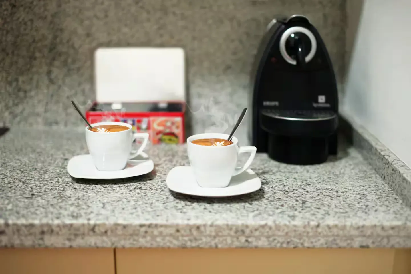 Dos tazas de café con cucharas, cafetera y caja de té sobre una encimera.