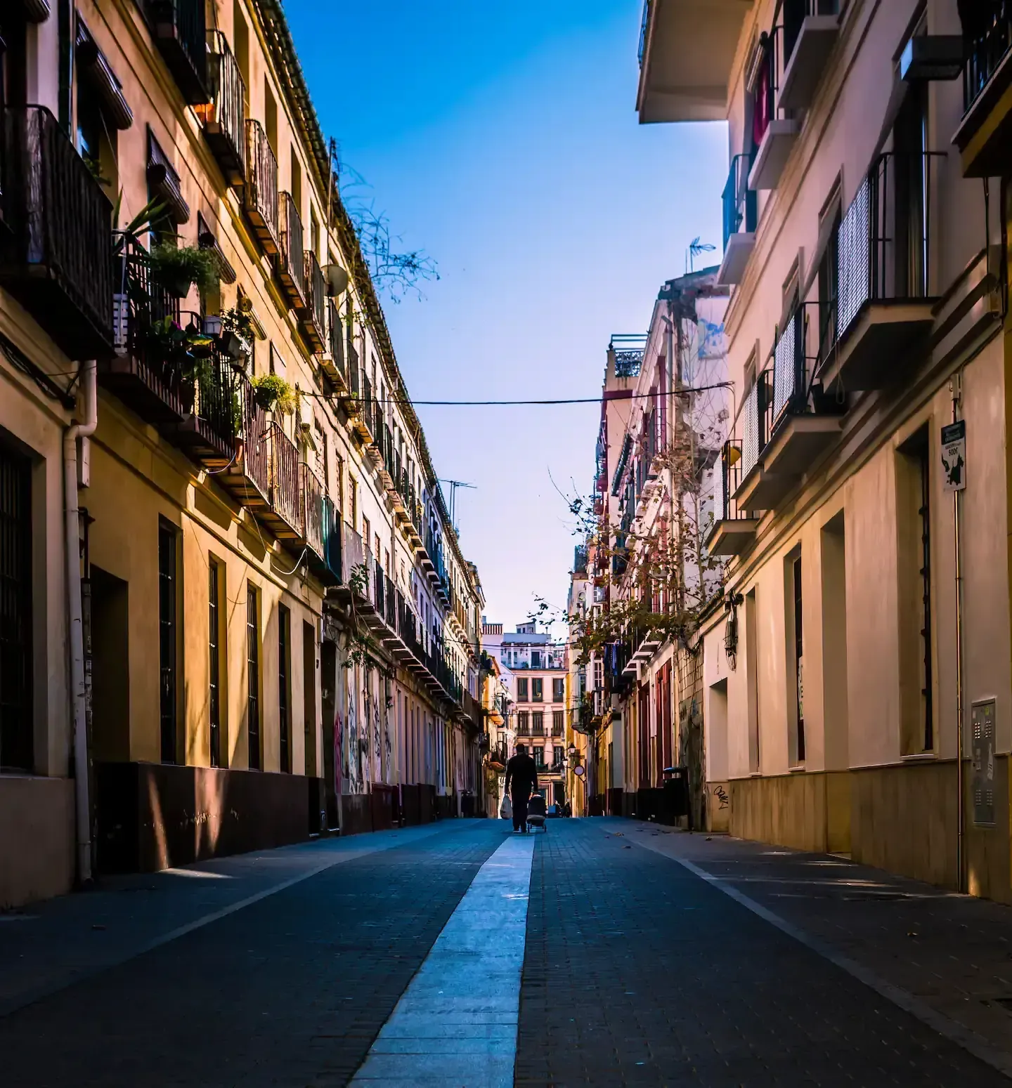 Calle estrecha con edificios beige, balcones y una figura que se aleja. Cielo azul.