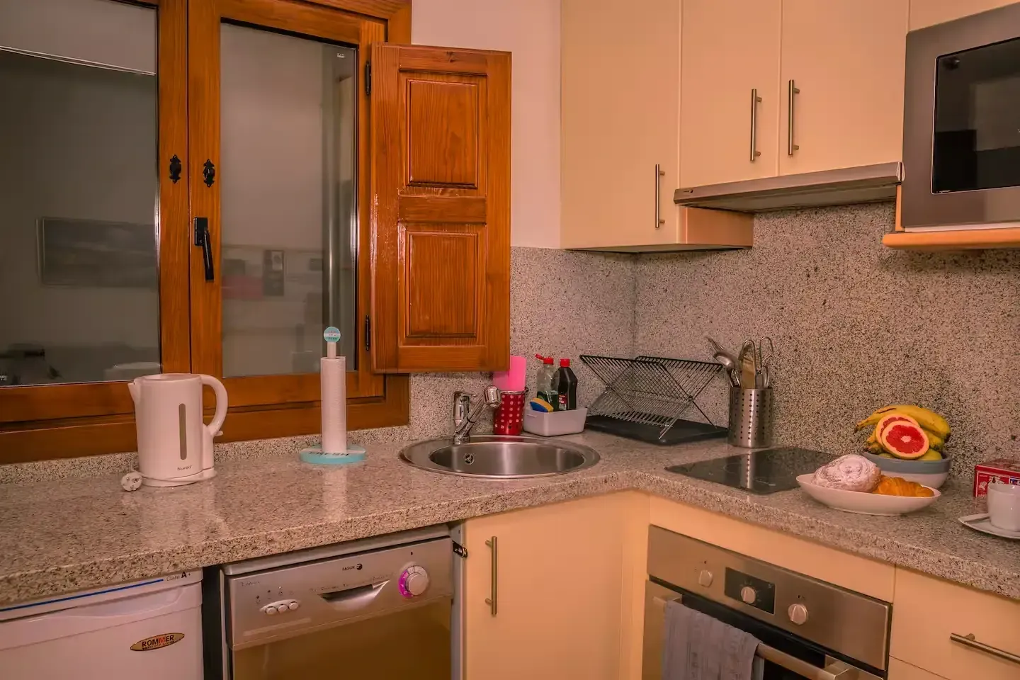 Una pequeña cocina con encimeras claras, gabinetes de color beige y una ventana con contraventanas de madera.