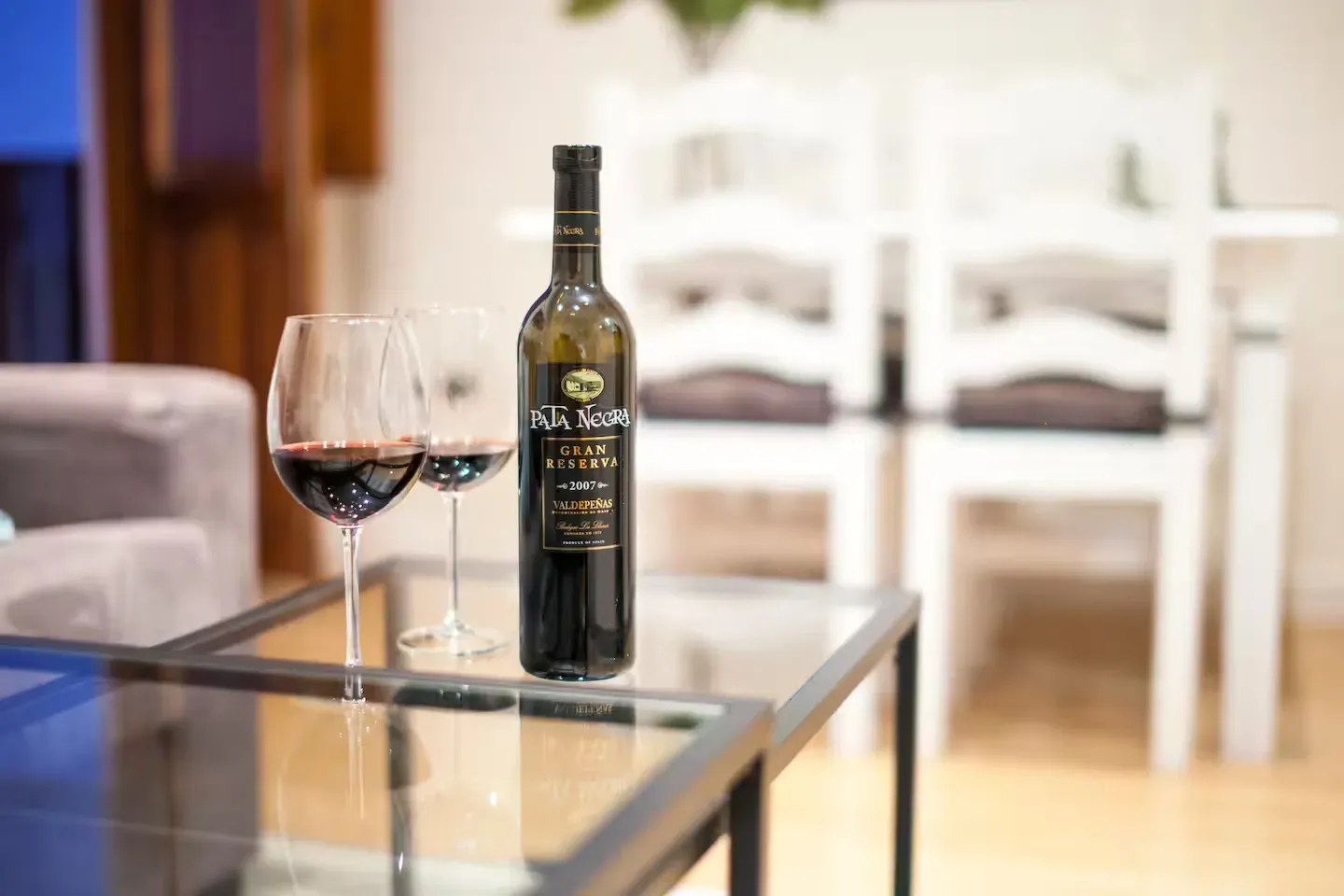 Botella de vino y copas sobre una mesa de cristal delante de un juego de comedor blanco.