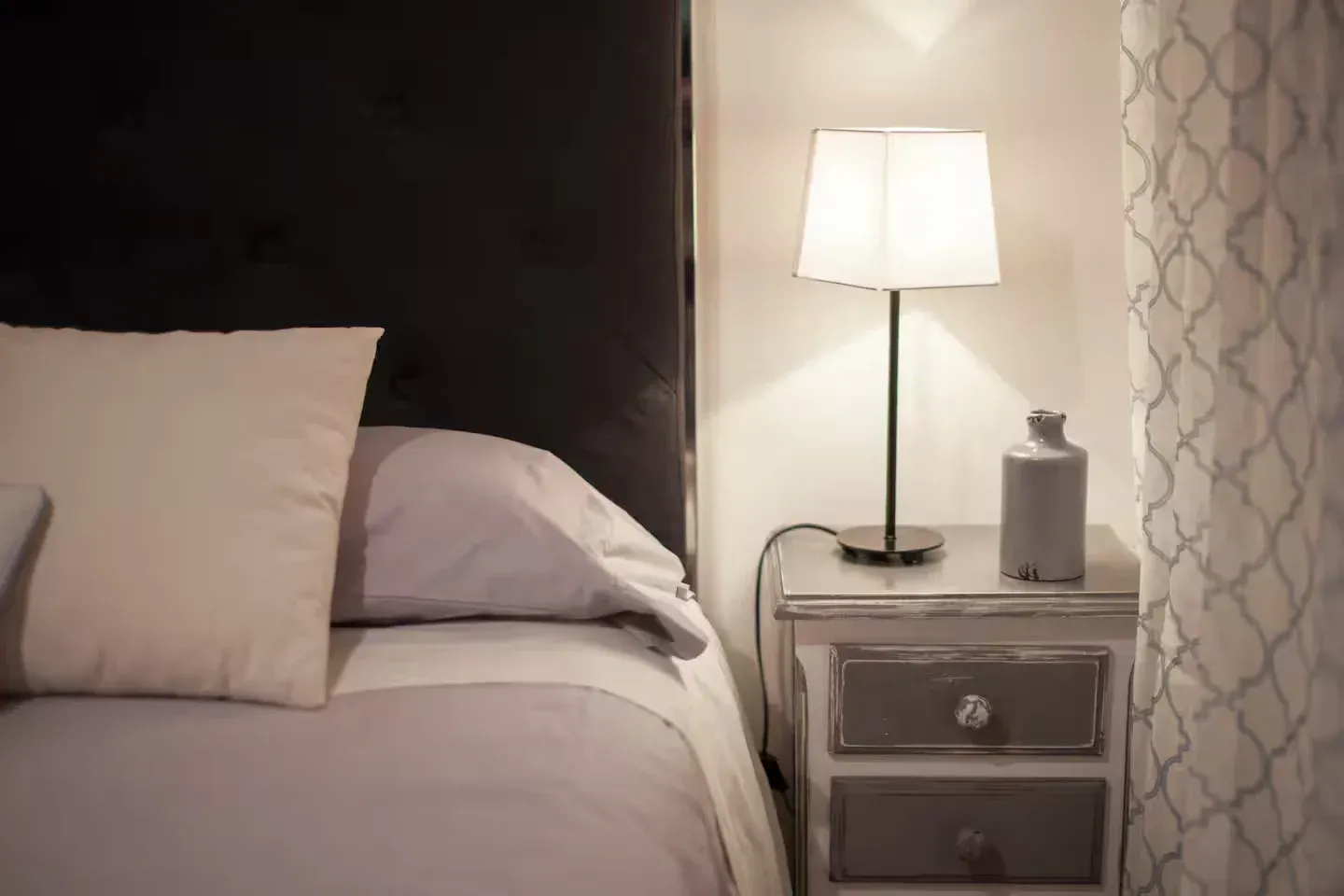 Mesita de noche con lámpara, almohadas y una botella decorativa.