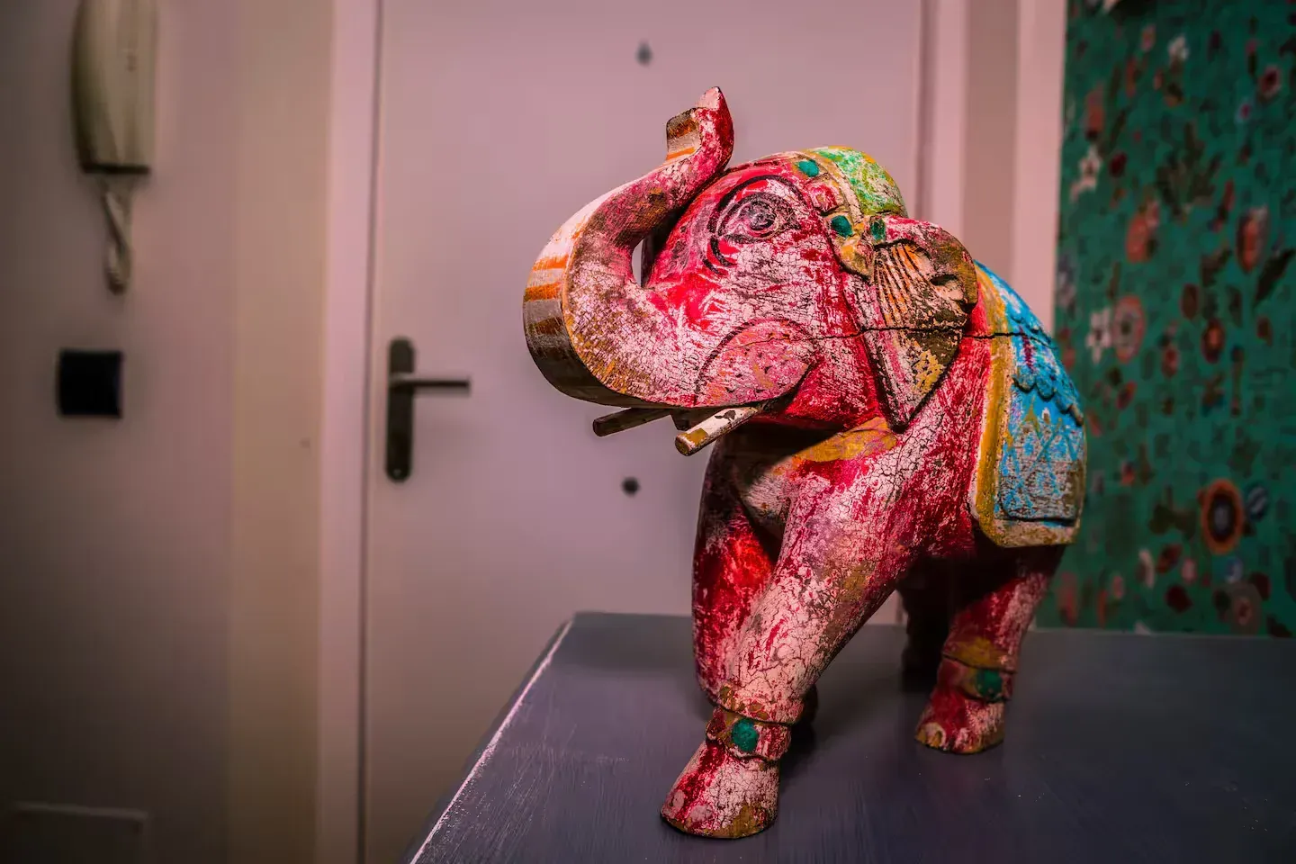 Estatua de elefante de madera colorida sobre una mesa, con una puerta y papel tapiz floral en el fondo.