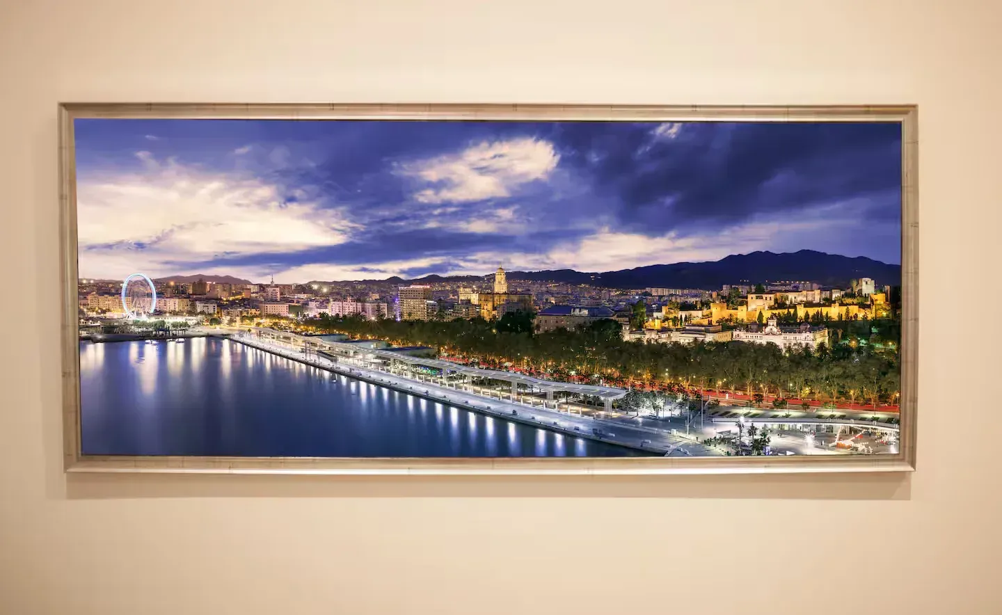 Paisaje urbano al anochecer, con luces reflejándose en el agua. Edificios, noria, montañas y un cielo nublado.