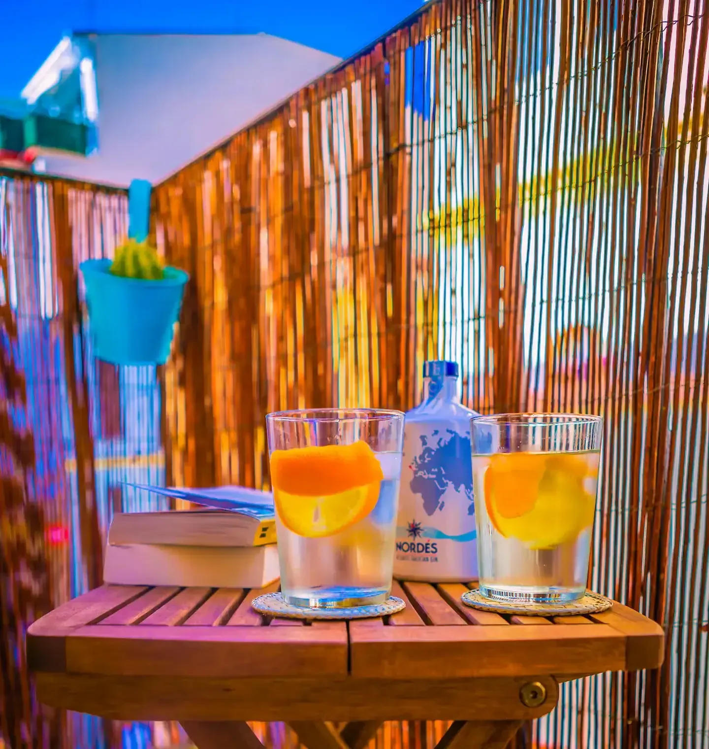 Dos bebidas con rodajas de naranja, una botella y un pequeño libro sobre una mesa de madera, contra una valla de bambú.