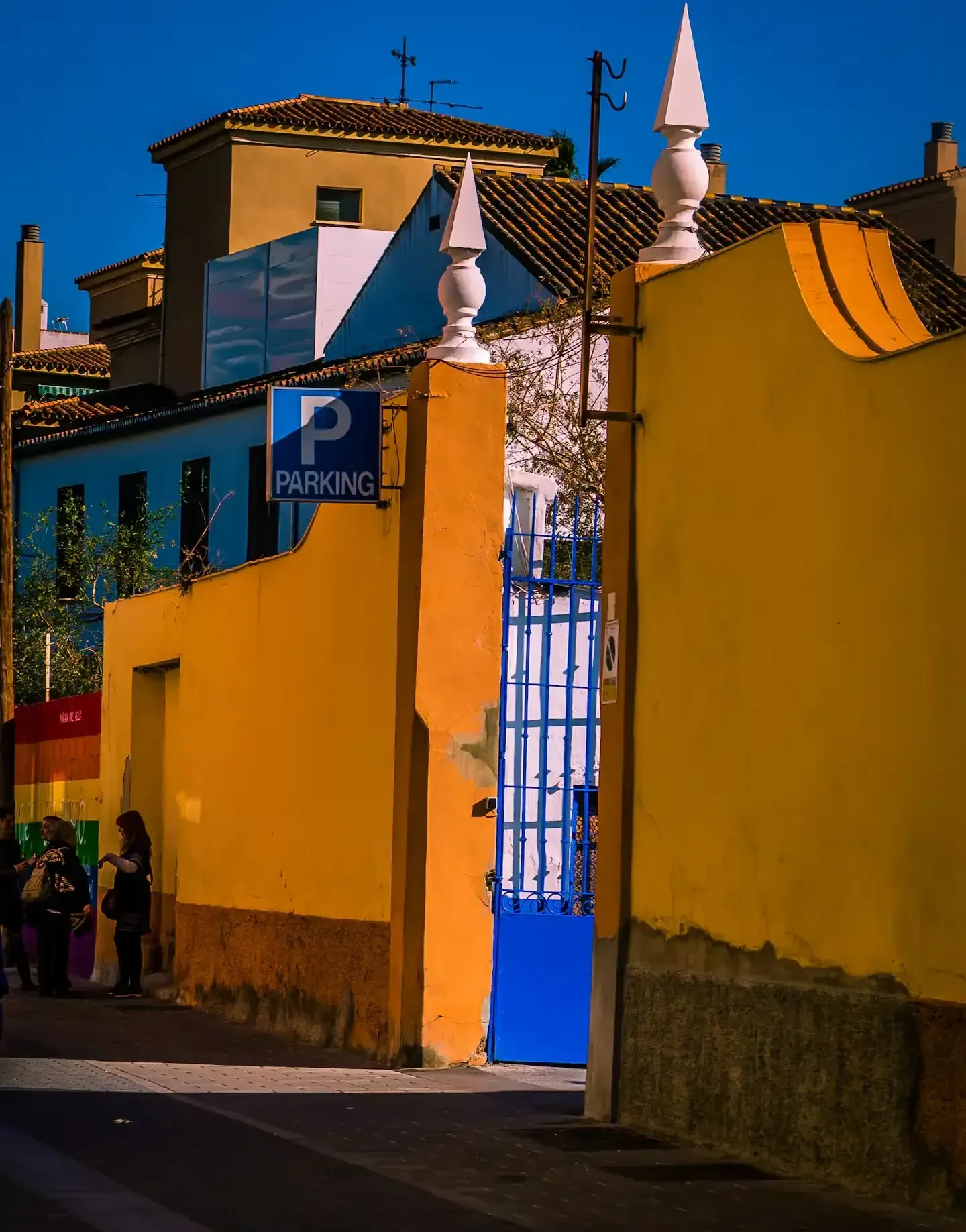Edificios brillantes enmarcan una puerta azul y un cartel de estacionamiento, con una bandera de arcoíris visible.