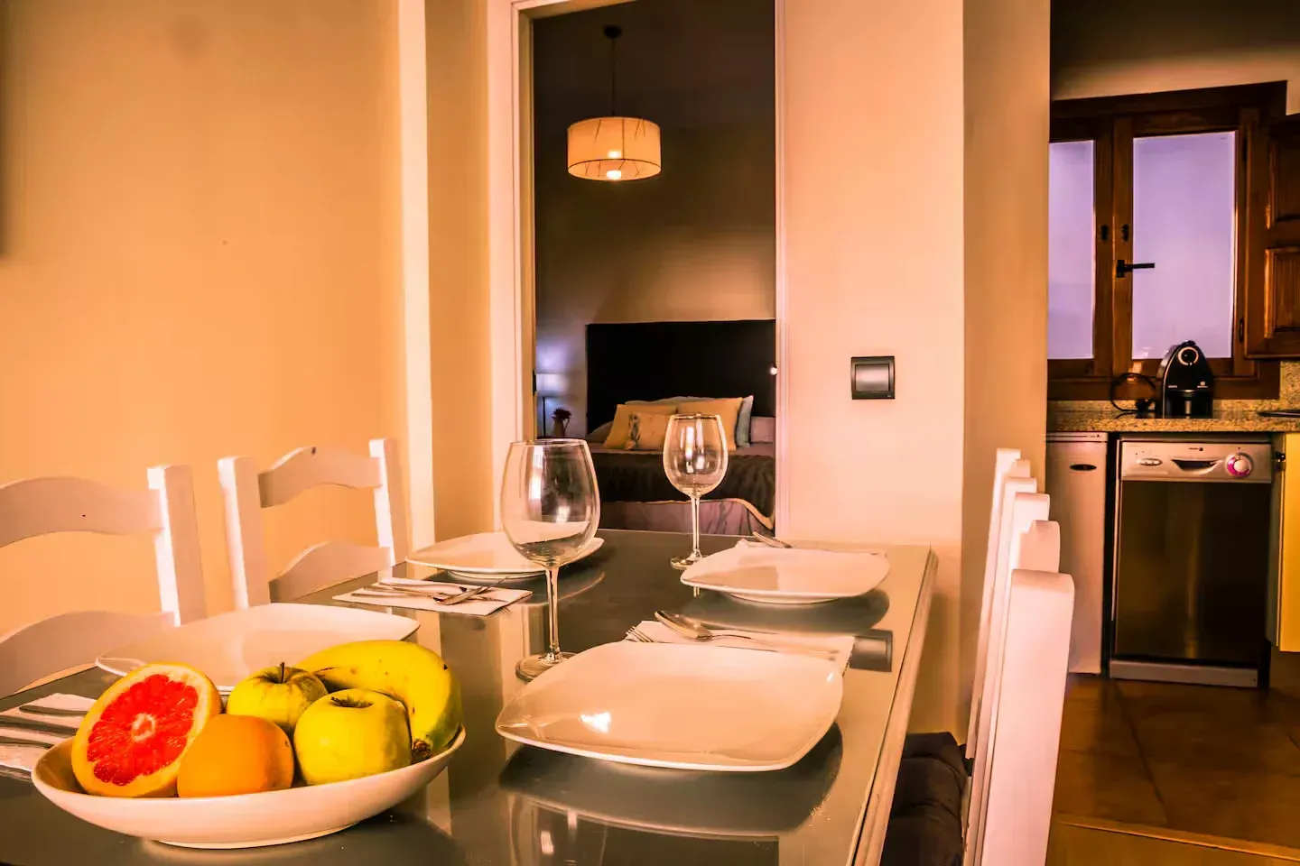 Mesa de comedor dispuesta para comer con frutero, vista de un dormitorio y cocina.