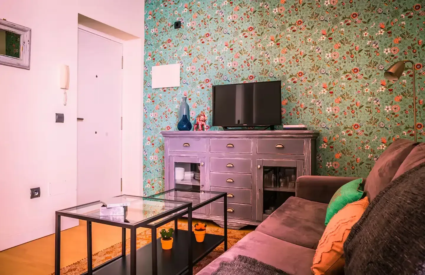 Sala de estar con papel tapiz floral, TV sobre un mueble gris, sofá marrón y mesa de centro de cristal.