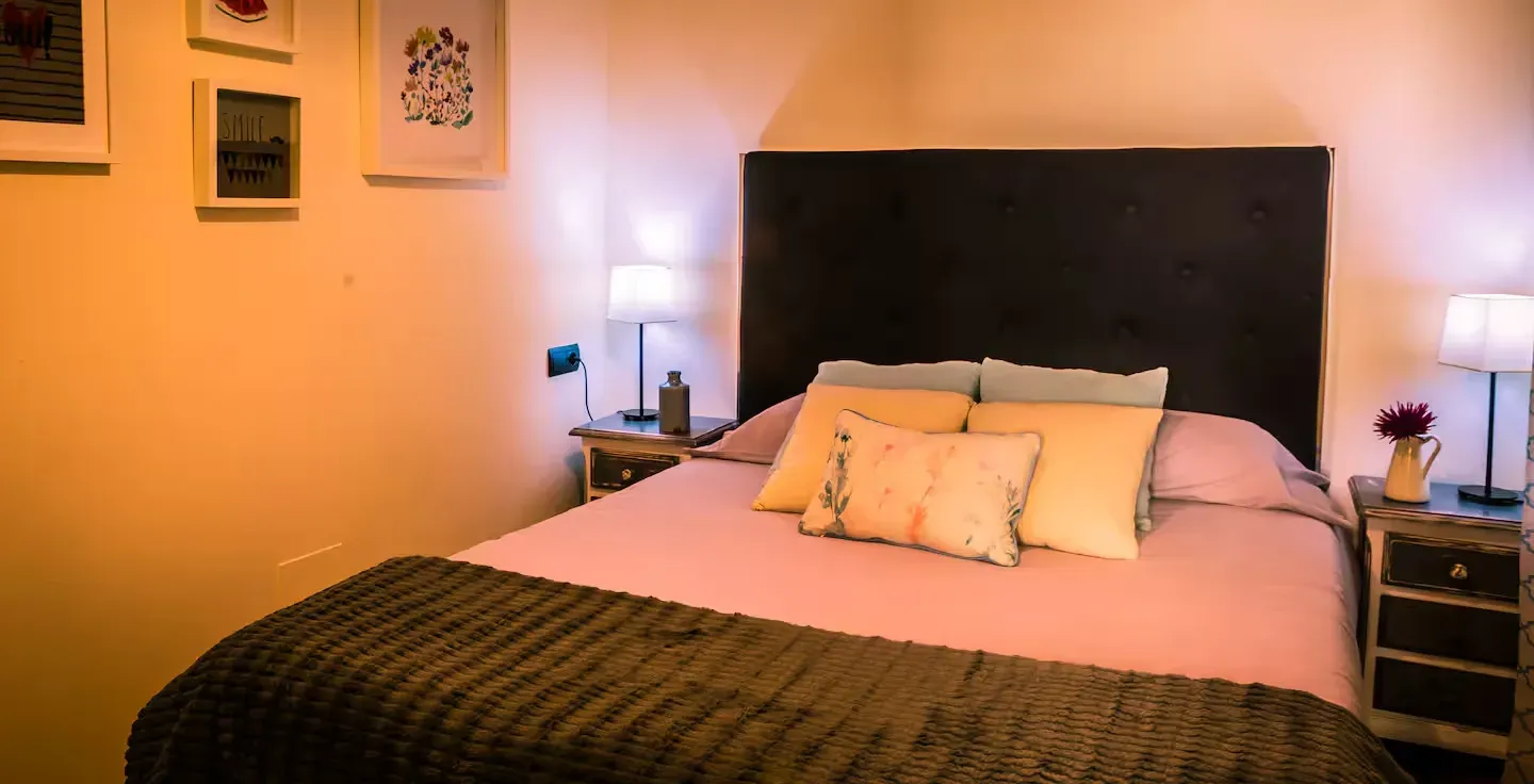 Dormitorio con colcha rosa, cabecero oscuro, lámparas pequeñas y arte enmarcado.