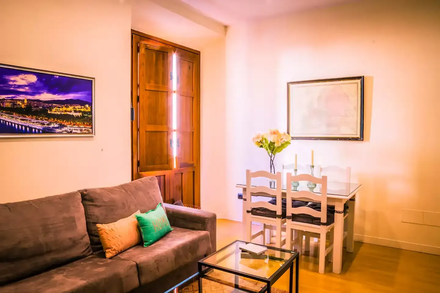 Sala de estar con un sofá gris, una pequeña mesa de cristal, una mesa y sillas blancas, arte y una puerta de madera.