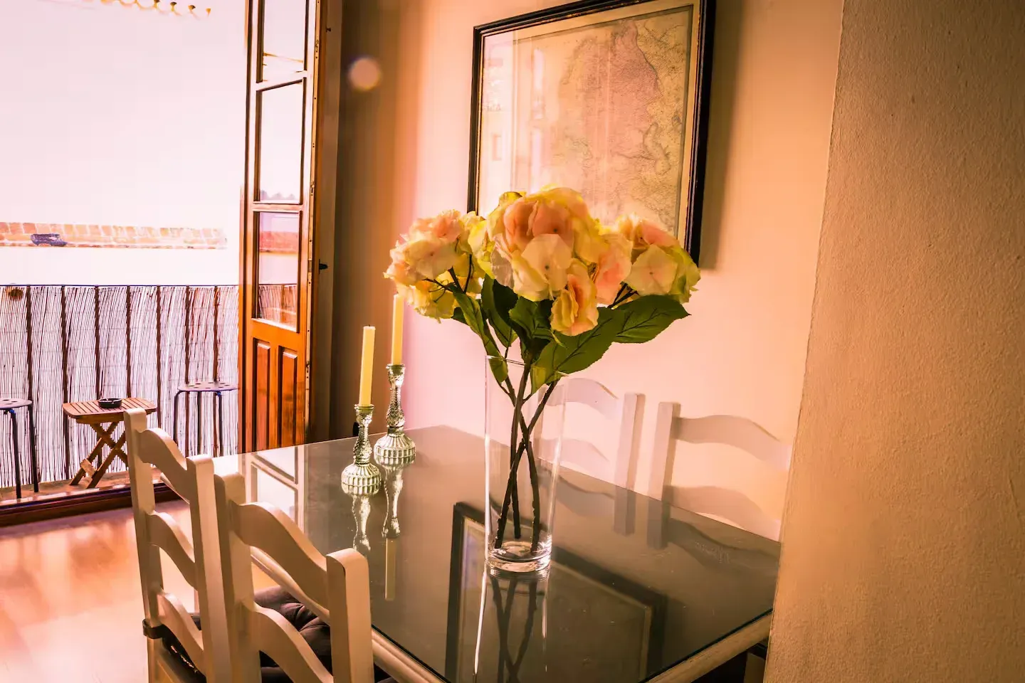 Mesa de comedor preparada para comer, con arreglo floral, puerta abierta al balcón.