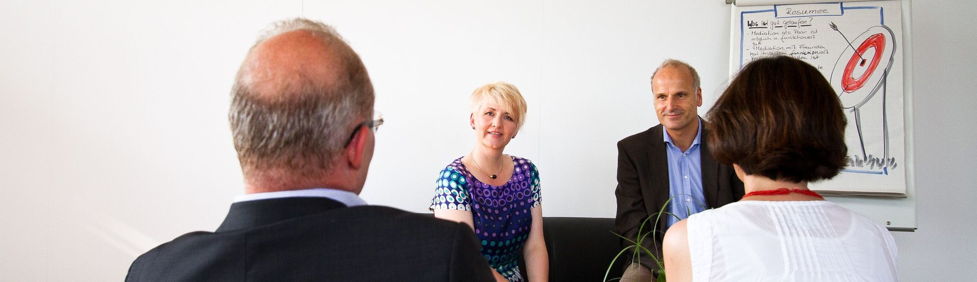 Mediation: Zwei Personen sitzen mit dem Rücken zur Kamera und blicken die Mediatorin und der Mediator an, die sitzen.