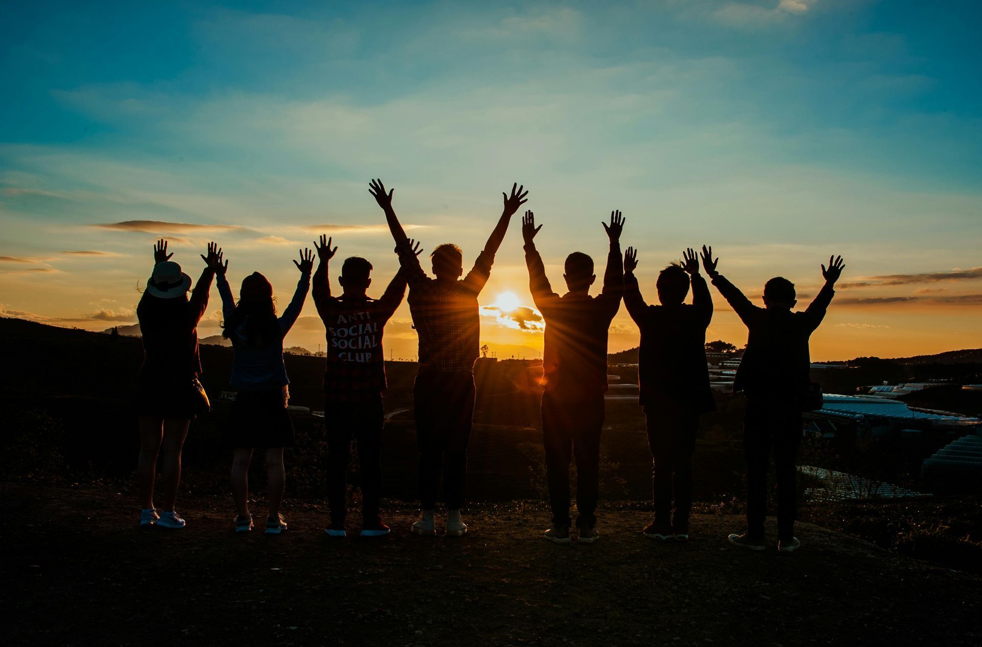 Eine Gruppe von sieben Personen steht als Silhouette mit erhobenen Armen vor einem hellen Sonnenuntergang.