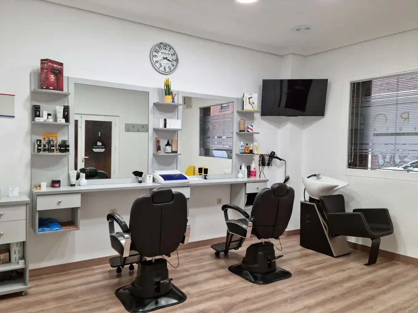 Interior de peluquería: sillas de barbero frente a espejos, productos en estantes, un reloj y un lavabo.