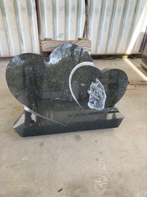 Pierre tombale en granit noir en forme de cœur avec des mains gravées, posée sur un sol en béton, sous un ciel couvert.