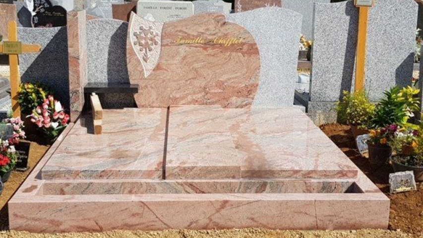 Pierre tombale en granit rose ornée d'un motif floral gravé. Située dans un cimetière avec des fleurs et d'autres pierres tombales.