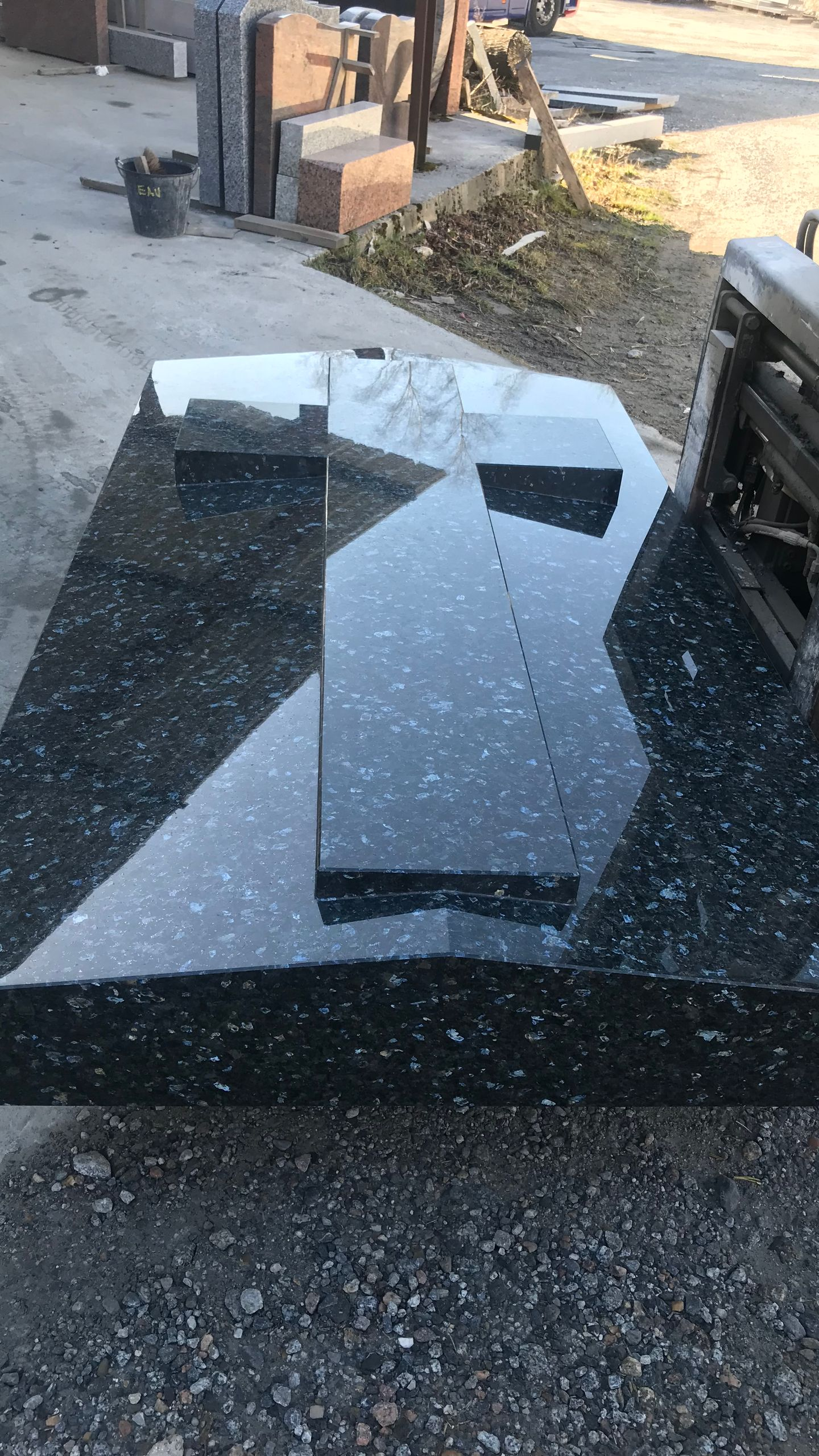 Pierre tombale en granit noir avec une croix posée dessus, à l'extérieur par une journée ensoleillée.