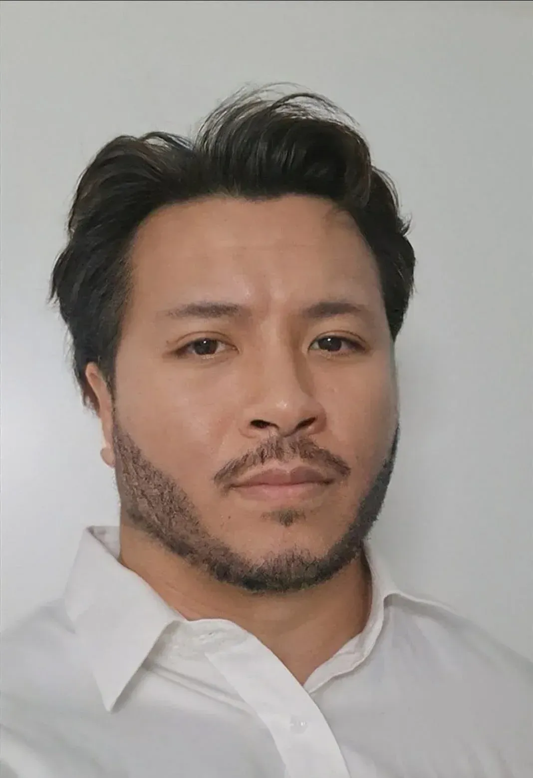 Tummatukkainen ja -partainen mies, jolla on valkoinen napitettava paita, katsoo suoraan kameraan.