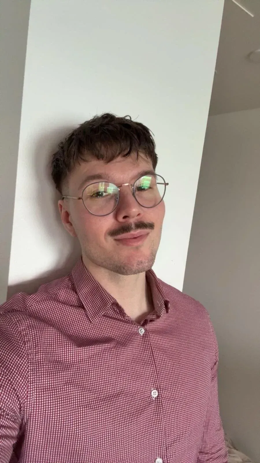 Silmälasit ja viikset omaava mies, jolla on yllään punavalkoruutupaita, nojaa valkoista seinää vasten ja hymyilee.