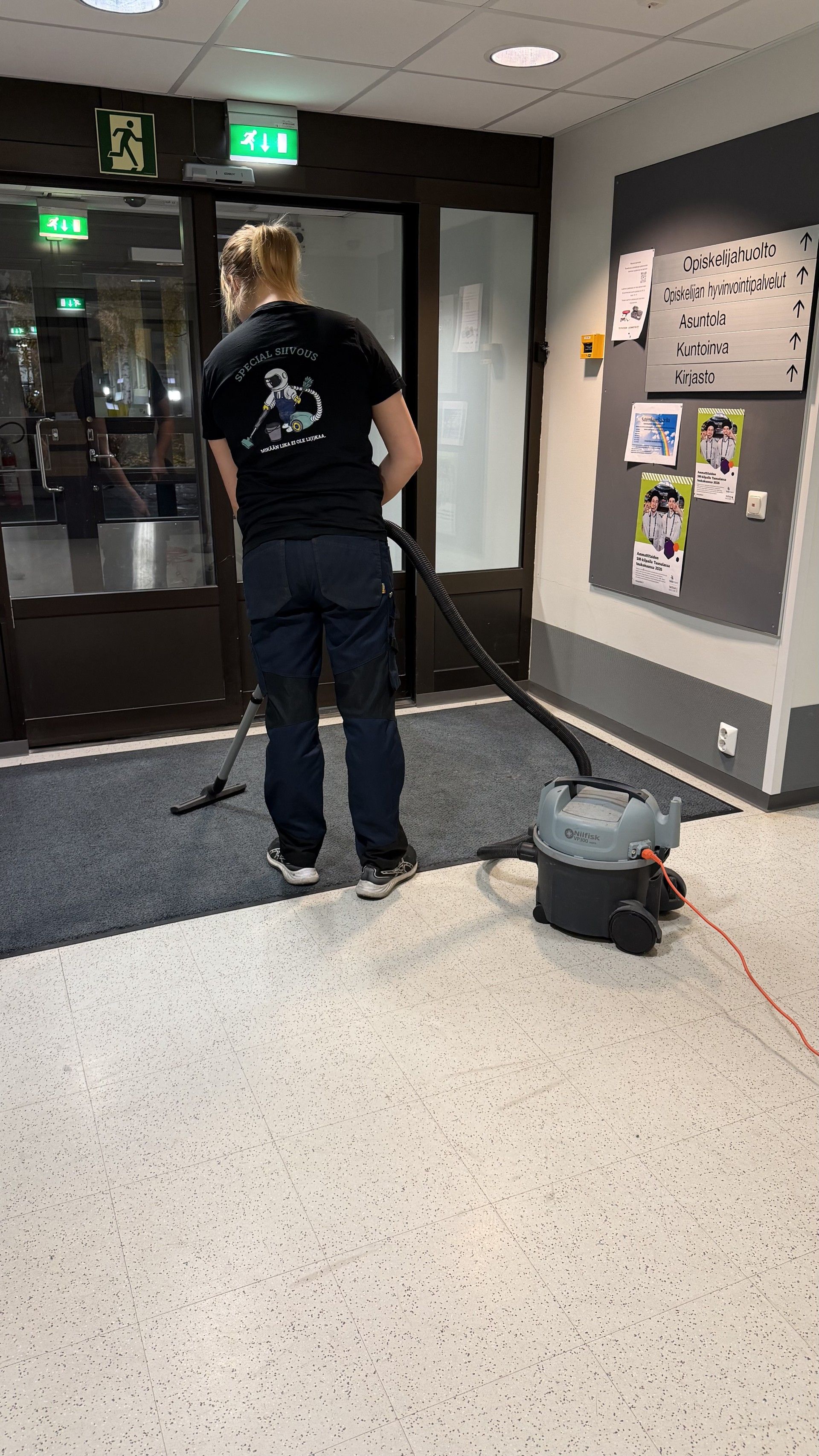 Henkilö imuroi tummaa mattoa rakennuksen sisällä oven ja ilmoitustaulun lähellä.