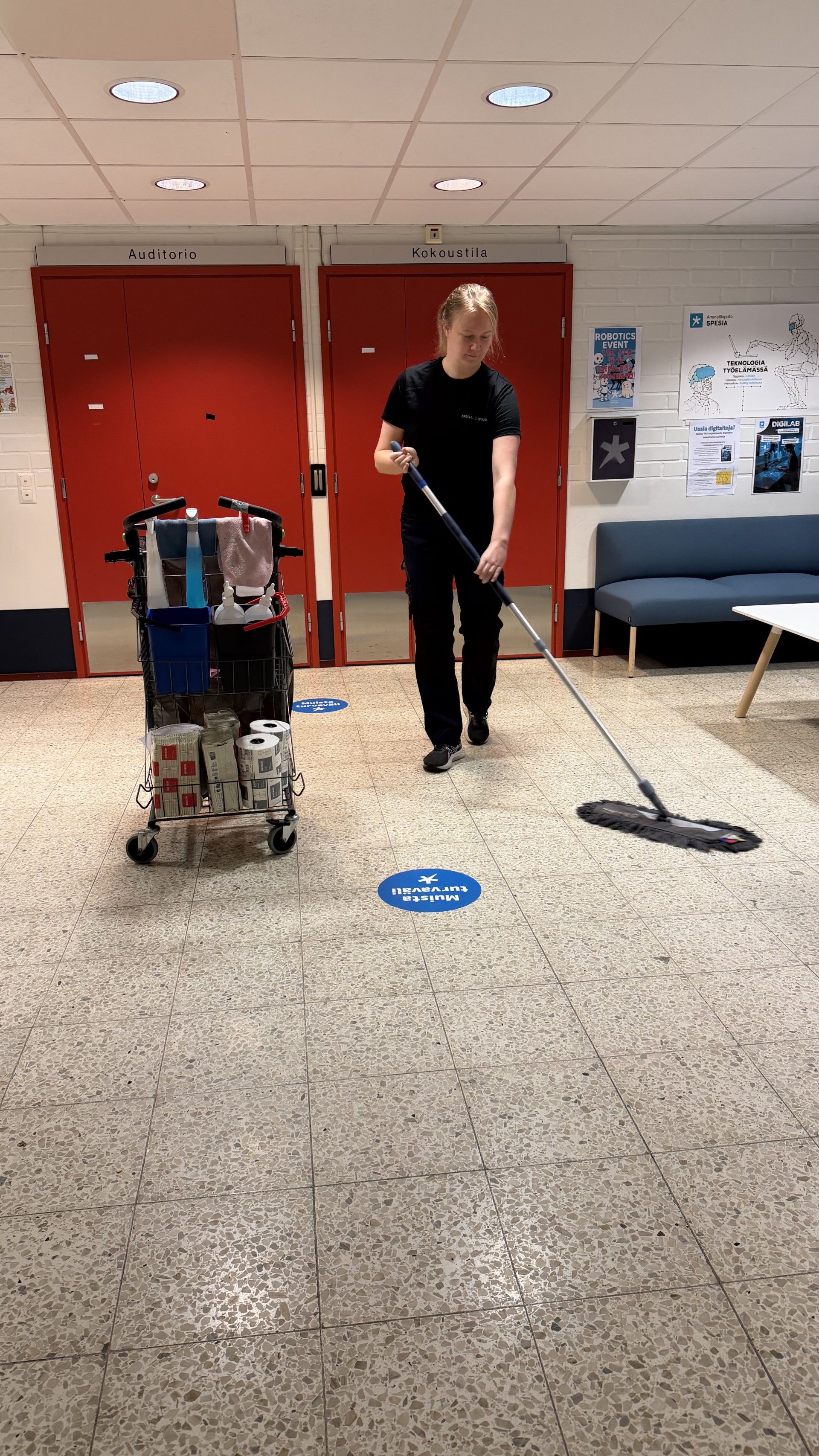 Henkilö moppaa lattiaa siivouskärryn vieressä punaovilla varustetussa käytävässä.