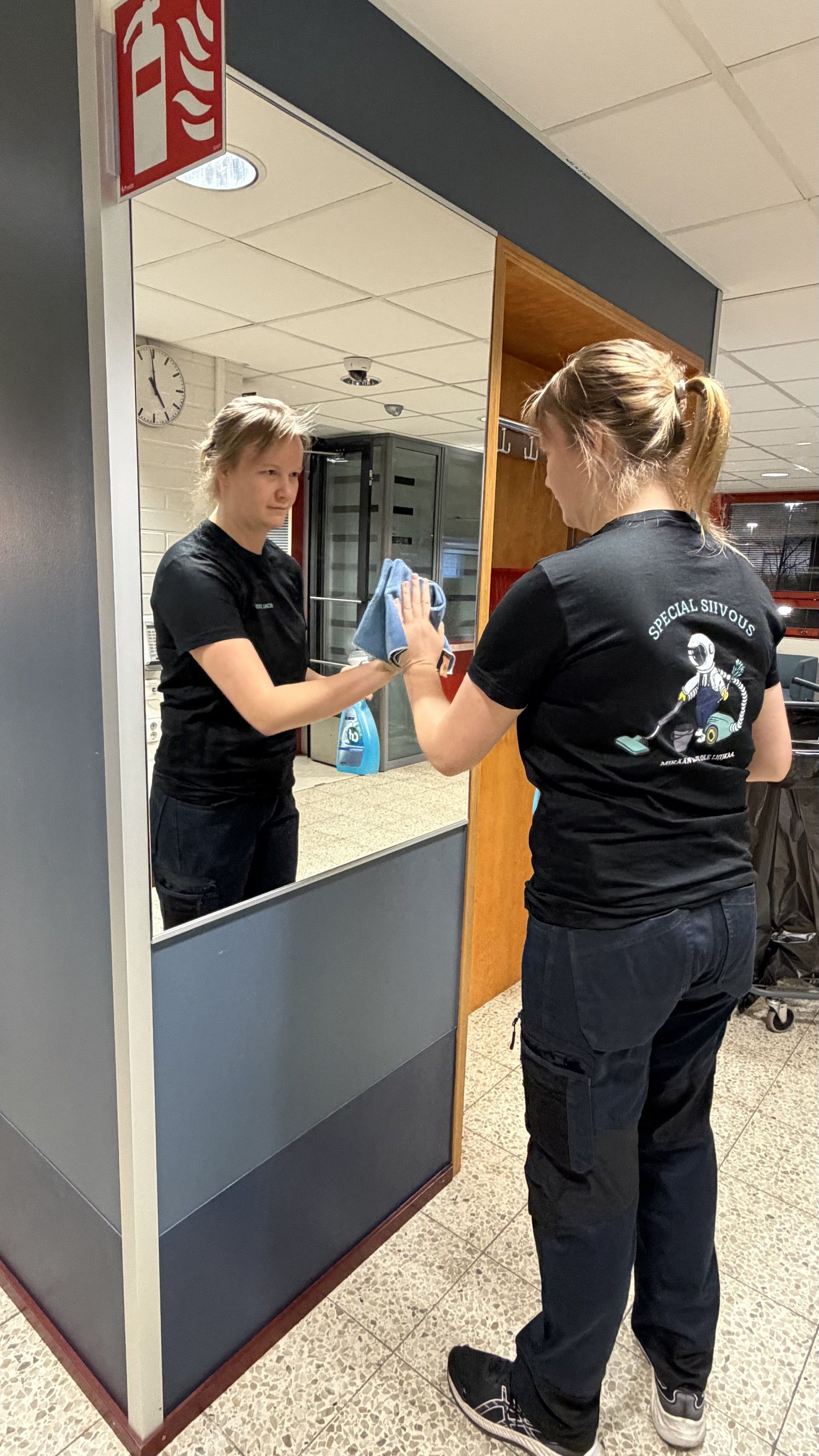 Henkilö puhdistaa peiliä käytävällä. Hänellä on yllään musta paita ja tummat housut, ja hän käyttää sinistä suihkepulloa ja liinaa.