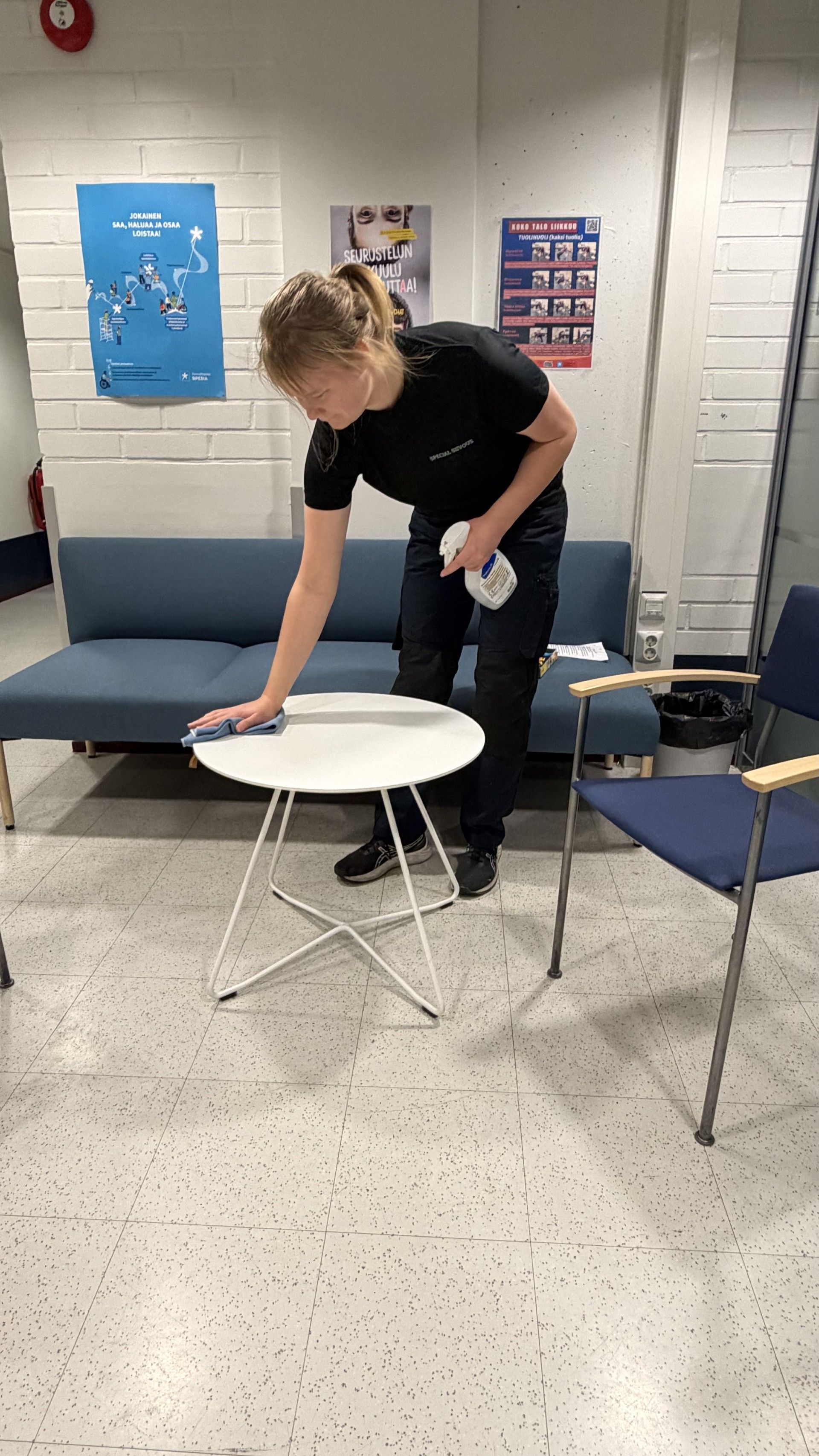 Henkilö puhdistaa pientä valkoista pöytää huoneessa, jossa on sinisiä huonekaluja ja julisteita seinillä.