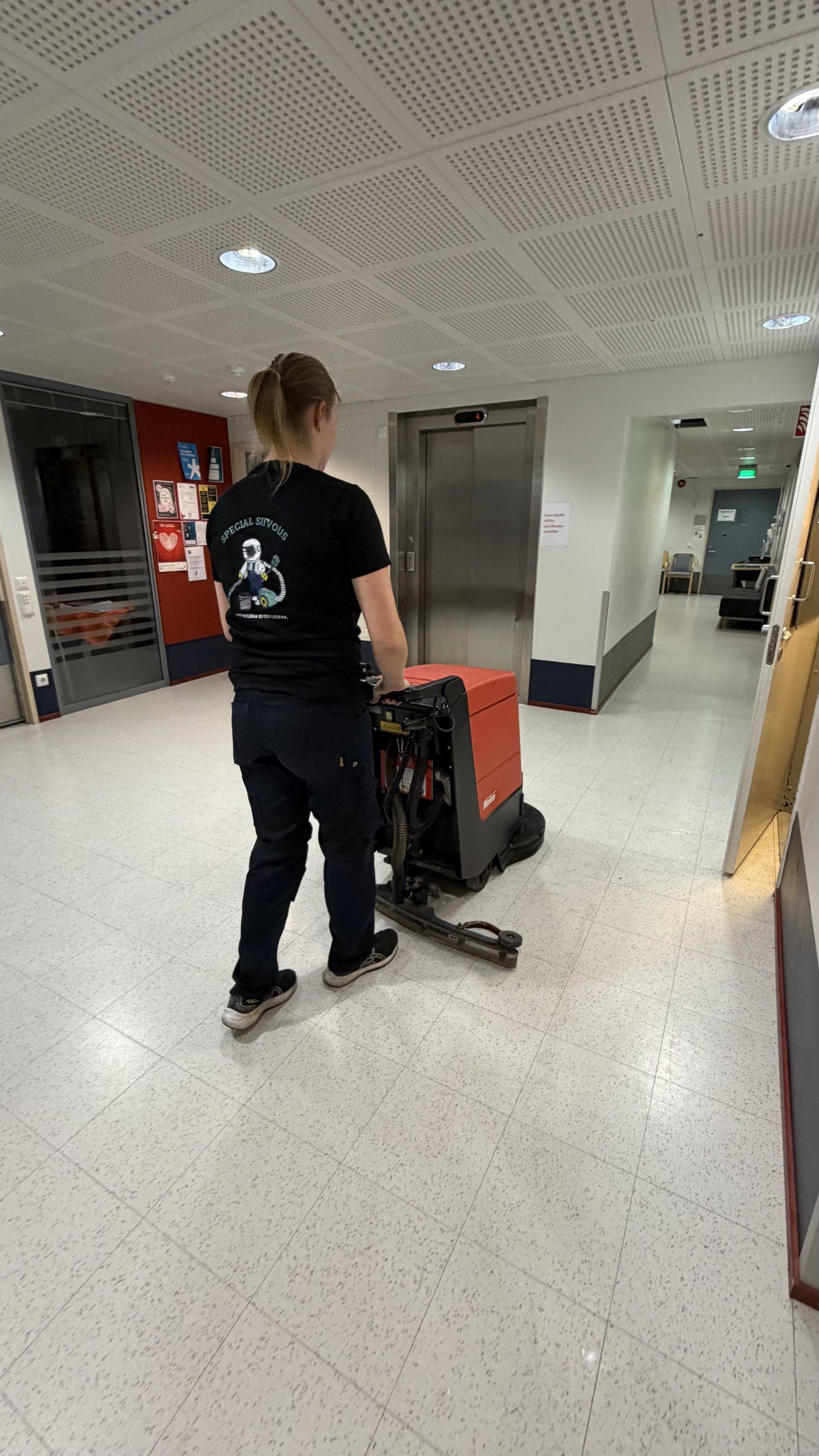 Henkilö käyttää lattianpesukonetta käytävällä, jossa on hissi ja julisteita.