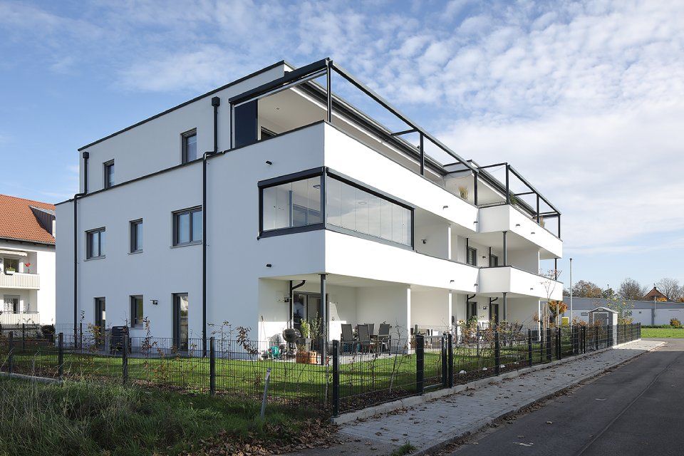 Modernes weißes Mehrfamilienhaus mit Balkonen zur Straße hin und umzäuntem Hof