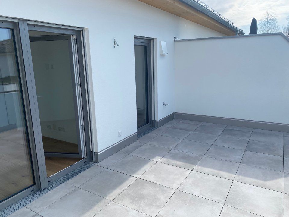 Kleine Dachterrasse mit grauen Fliesen, weißen Wänden und Glasschiebetüren zum Innenraum.