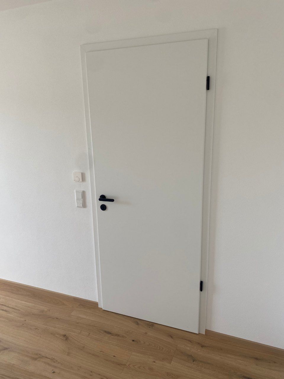 Weiße Innentür mit schwarzem Türgriff in einem schlichten Raum mit Holzboden.