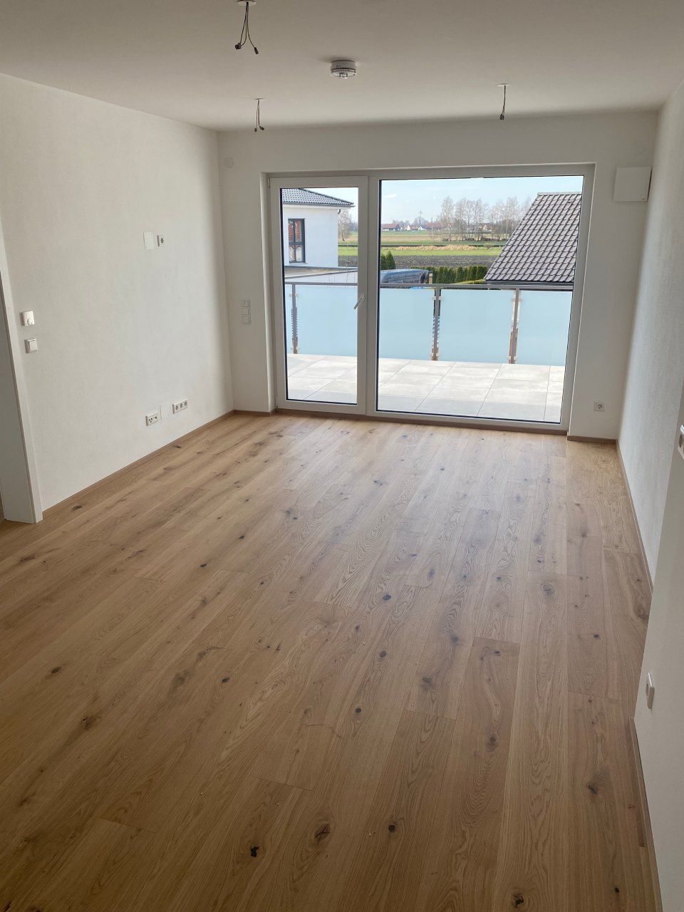 Leeres, unmöbliertes Zimmer mit Holzboden und Glasschiebetüren, die auf einen Balkon mit Wasserblick führen.