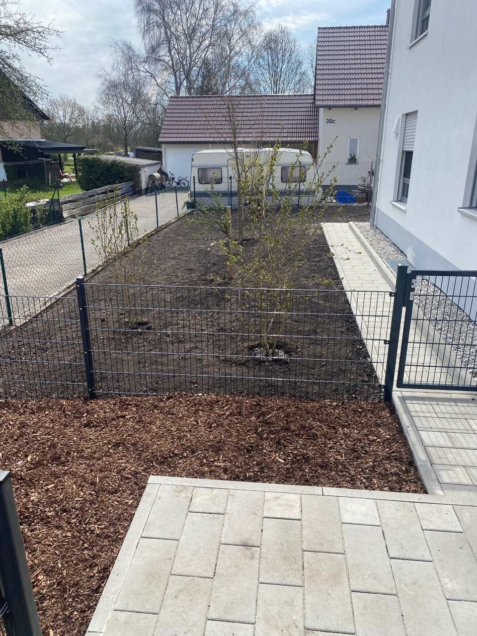 Gartenbeet im Hinterhof mit frischer Erde neben einem weißen Haus und einer gepflasterten Terrasse, beidseitig eingezäunt