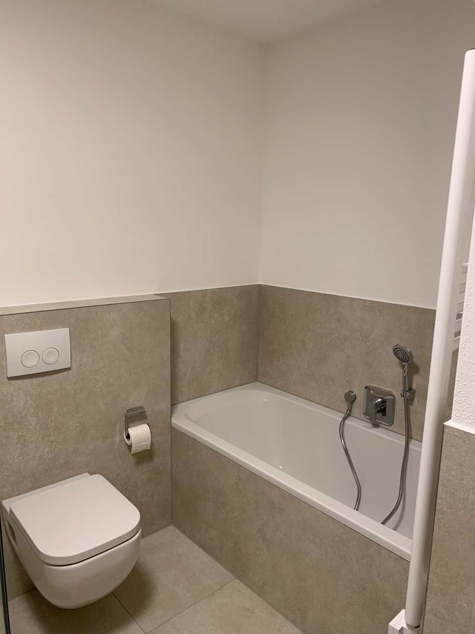 Kleines Badezimmer mit wandhängendem WC, Badewanne und beigefarbener Fliesenvertäfelung.