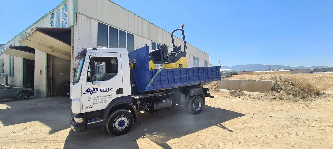 Un camión blanco con una caja azul que transporta equipos de construcción cerca de un edificio en un día soleado.