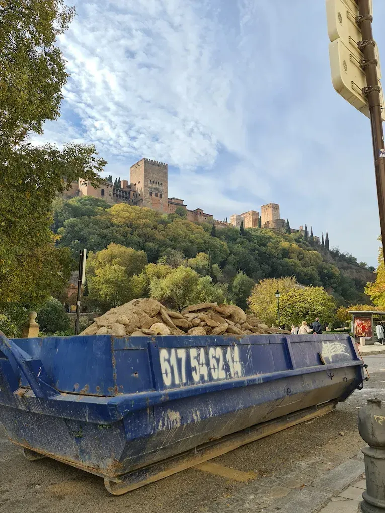 Contenedor azul lleno de escombros en una calle, con el castillo de la Alhambra en una ladera verde al fondo.