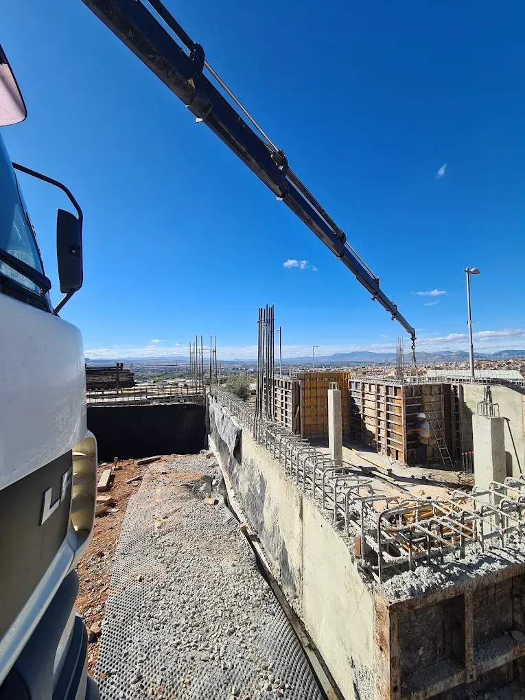 Sitio de construcción con encofrados de hormigón, barras de refuerzo y una pluma de grúa contra un cielo azul.