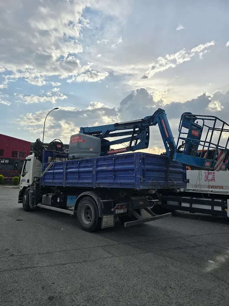 Camión azul con grúa cargando un remolque bajo un cielo nublado.