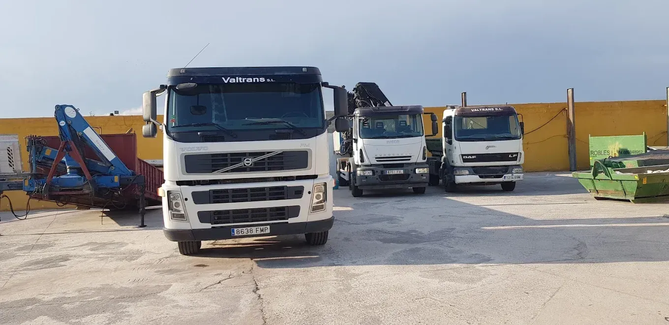 Tres camiones de trabajo estacionados en un estacionamiento: un Volvo al frente, camiones blancos detrás. Cielo brillante, pared amarilla.