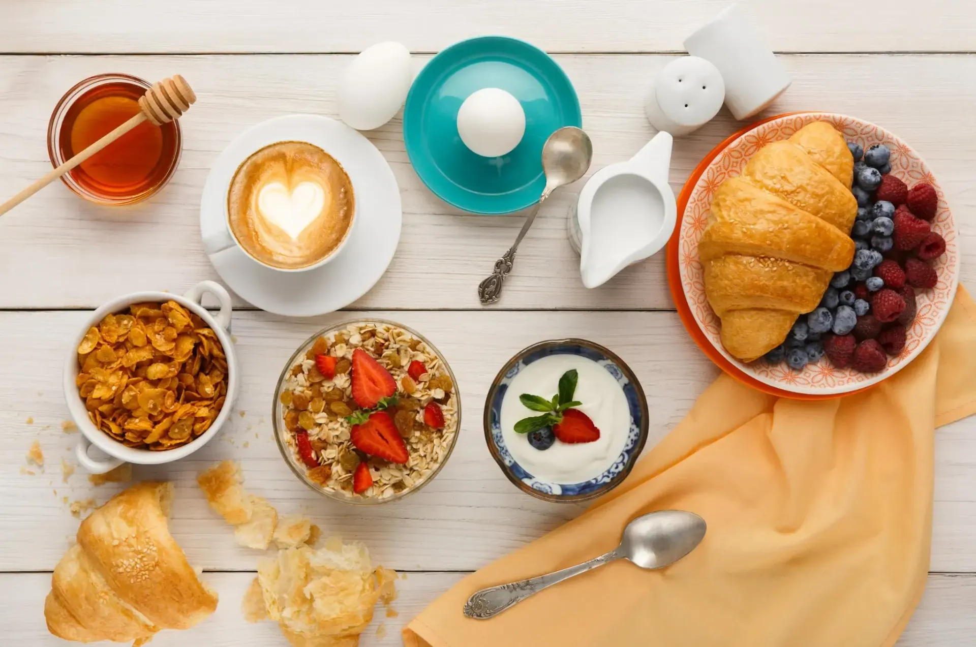 Desayuno servido en una mesa de madera blanca: café, granola, avena, yogur, croissant con frutos rojos, huevo cocido y miel.