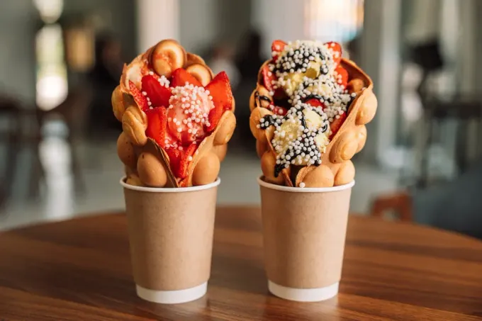 Dos waffles de burbujas rellenos de fresas y aderezos, servidos en vasos de papel, sobre una mesa de madera.