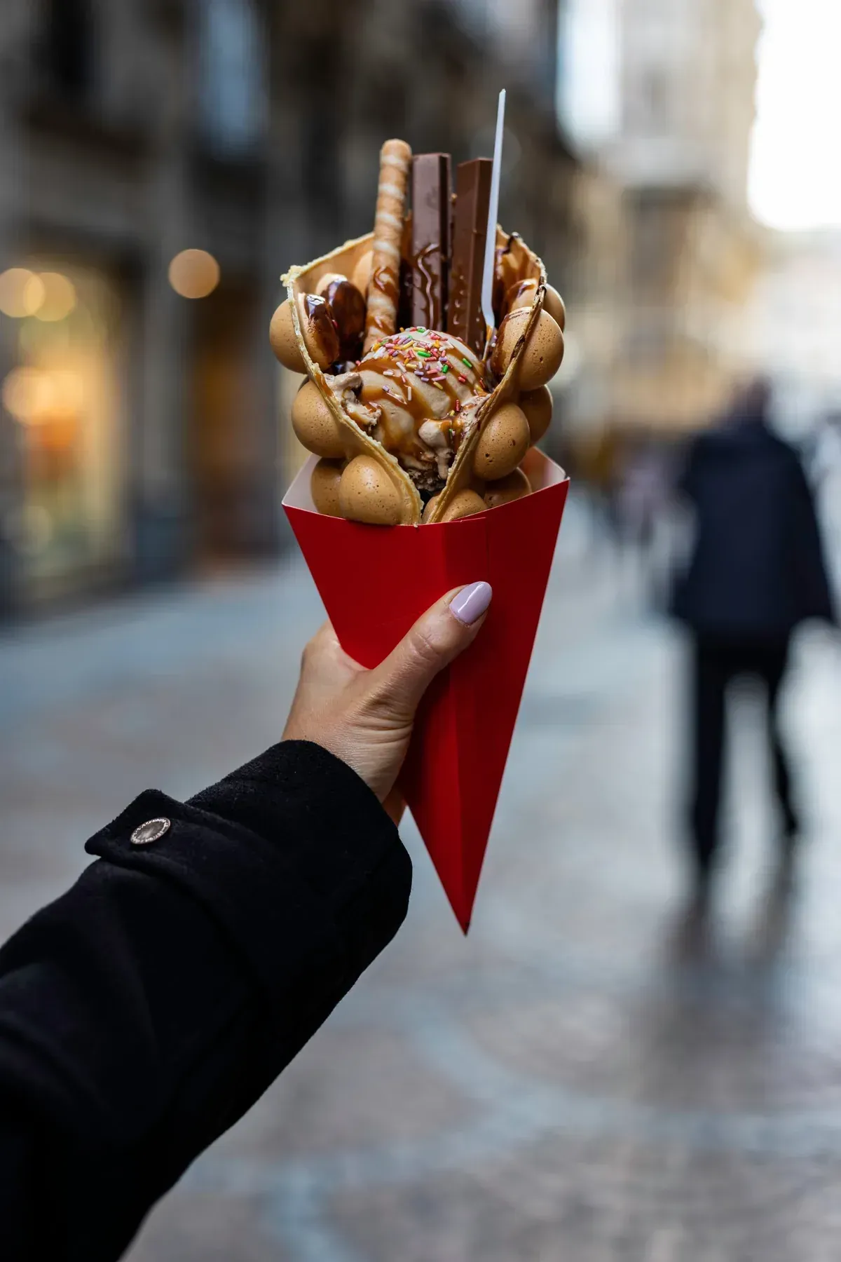 Mano sosteniendo un cono de papel rojo lleno de un waffle de burbujas y aderezos, en una calle de la ciudad.