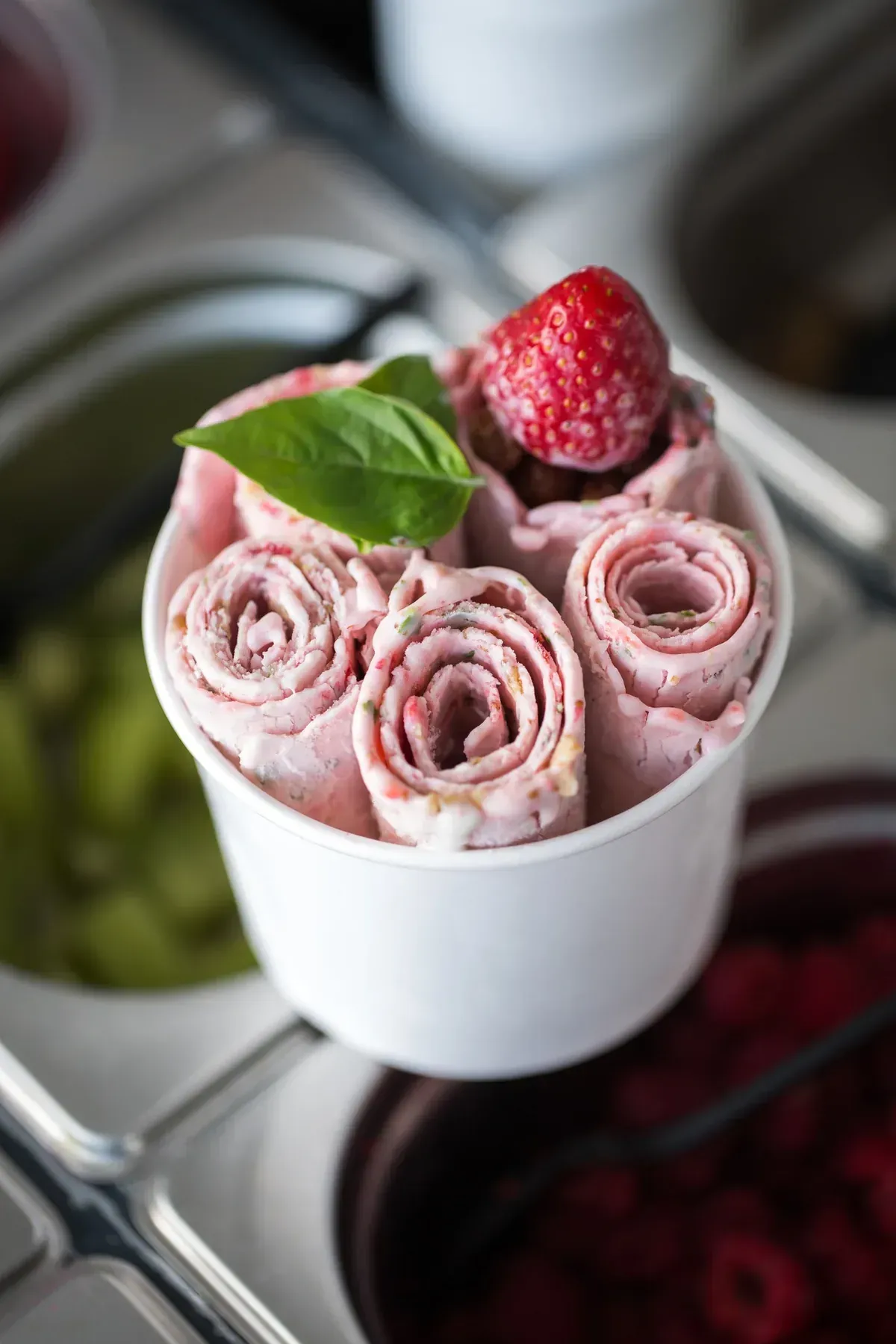 Rollos de helado en vaso blanco, con fresa y albahaca. Rosa, con ingredientes al fondo.