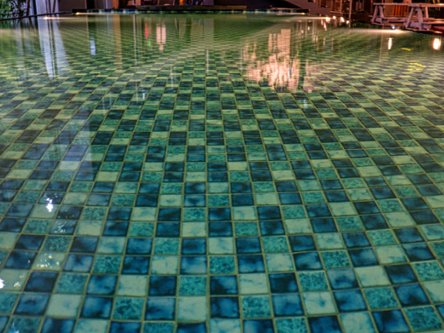 Piscine carrelée en damier reflétant les lumières et les bâtiments ; carrés turquoise et bleu foncé.