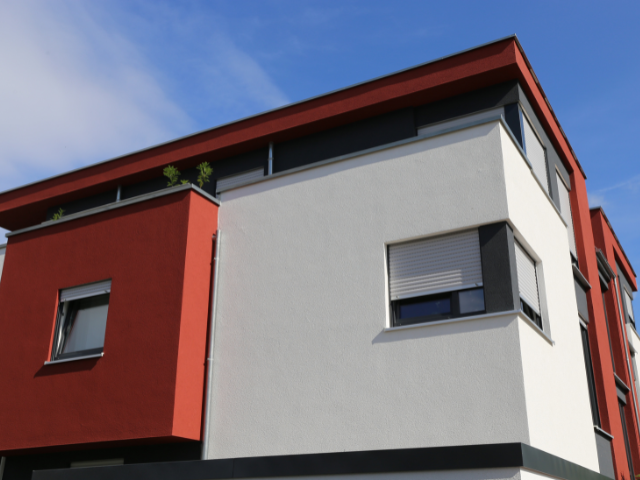 Bâtiment moderne de deux étages à la façade rouge et blanche. Stores fermés, ciel bleu en arrière-plan.