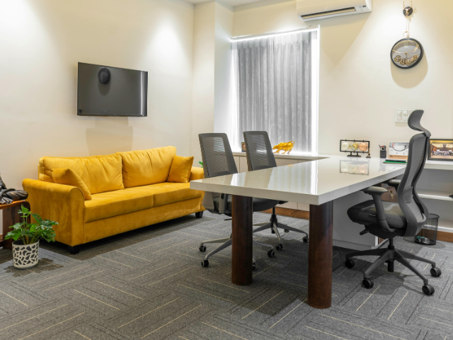 Bureau avec canapé jaune, grand bureau et chaises noires. Téléviseur au mur.