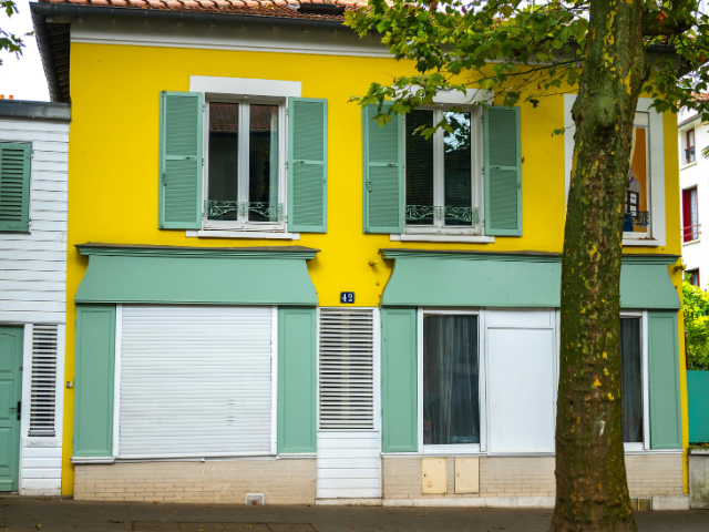 Maison jaune avec volets et auvents verts, arbre devant.