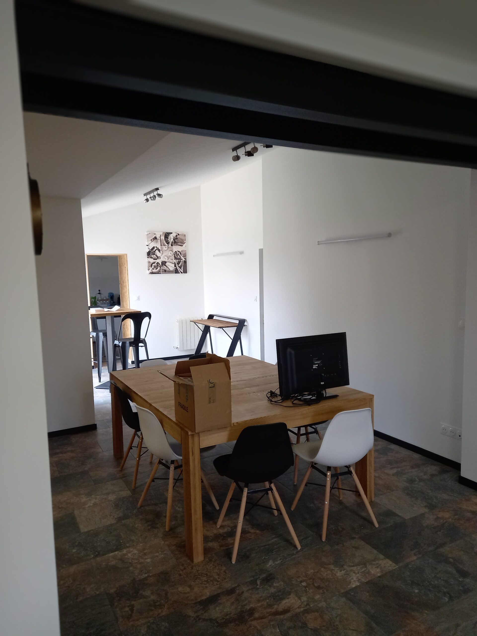 Salle à manger avec table en bois, chaises mixtes et un moniteur.