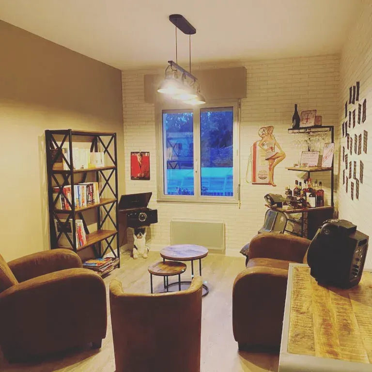 Salon chaleureux avec fauteuils marron, bibliothèque, petites tables et fenêtre. Éclairage chaleureux.