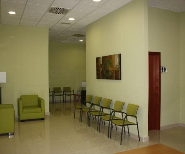 Sala de espera vacía con sillas verdes, un cuadro en la pared y una puerta roja.