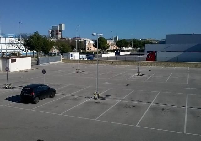 Estacionamiento vacío con un auto oscuro, postes de luz altos y edificios al fondo.